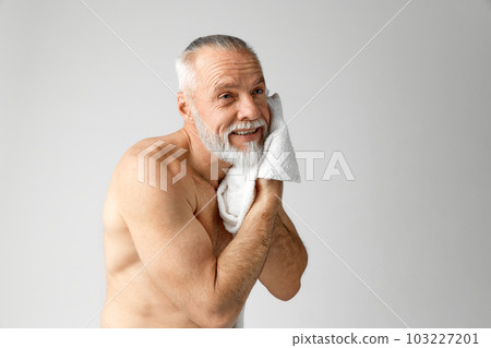 Portrait of handsome, mature, grey-haired, shirtless man doing morning routine, washing face against grey background 103227201