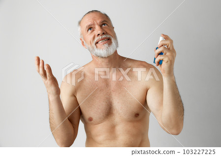 Portrait of handsome, smiling, mature man with gray hair and beard posing shirtless, applying with face toner against grey background Portrait of handsome, smiling, mature man with gray hair and beard posing shirtless, applying with face toner against grey background 103227225
