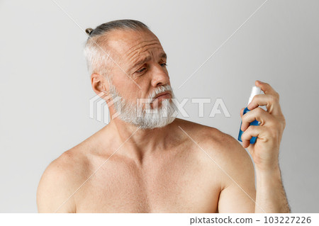 Portrait of serious, handsome, mature man with gray hair and beard posing with face toner against grey background Portrait of serious, handsome, mature man with gray hair and beard posing with face toner against grey background 103227226
