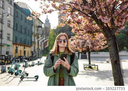Happy stylish clothes blond girl tourist travels looking for directions on smartphone while traveling through the old town of Krakow near the Wawel Royal Castle in spring, Poland. Happy stylish clothes blond girl tourist travels looking for directions on smartphone while traveling through the old town of Krakow near the Wawel Royal Castle in spring, Poland. 103227733