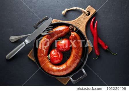 Fried pork sausage in a cast iron pan with tomatoes, hot peppers and cutlery on a wooden cutting board on a dark stone table Fried pork sausage in a cast iron pan with tomatoes, hot peppers and cutlery on a wooden cutting board on a dark stone table 103229406