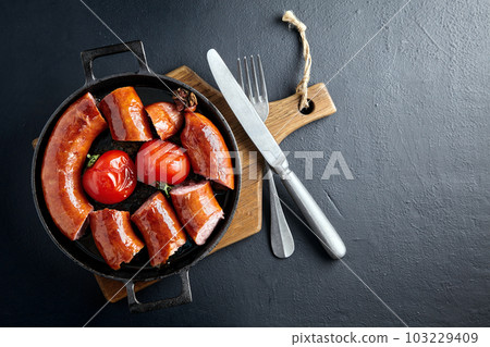 Portion cut fried pork sausage in a cast iron pan with tomatoes and cutlery on a wooden cutting board on a dark stone table 103229409