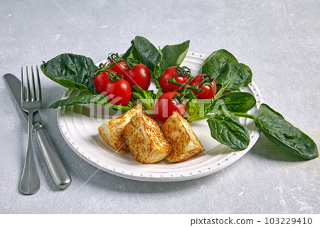 Cherry tomatoes on a branch with spinach and fried halloumi cheese on a ceramic gray plate on a light stone table 103229410