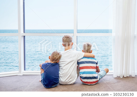 Eldest boy hugs younger brothers sitting on floor and looking at azure sea from panoramic window. Siblings enjoy spending summer holidays together Eldest boy hugs younger brothers sitting on floor and looking at azure sea from panoramic window. Siblings enjoy spending summer holidays together 103230130