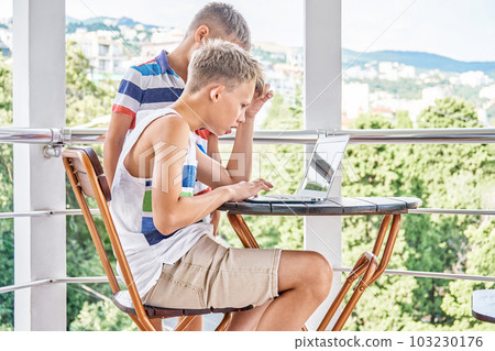 Brother siblings look together at difficult game on grey laptop on hotel balcony. Preteen schoolboy tries to win serious game during summer holidays Brother siblings look together at difficult game on grey laptop on hotel balcony. Preteen schoolboy tries to win serious game during summer holidays 103230176