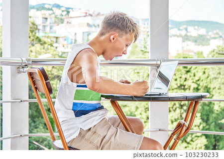 Blond schoolboy celebrates victory playing difficult video game on laptop. Preteen boy plays game spending summer holidays on hotel balcony 103230233