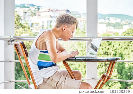 Preteen boy sitting on hotel balcony and surfing the internet on grey laptop. Schoolboy enjoys spending summer holidays and playing games Preteen boy sitting on hotel balcony and surfing the internet on grey laptop. Schoolboy enjoys spending summer holidays and playing games 103230299