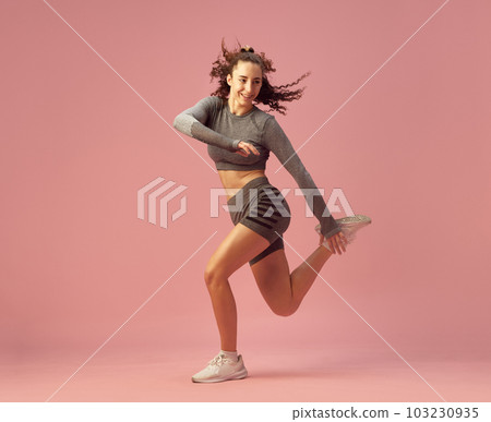 Dynamic image of sportive young girl in gray sportswear training, jumping against pink studio background 103230935
