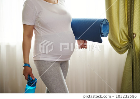 Close-up view of the belly of pregnant woman in 3d trimester of pregnancy, wearing a white mockup t-shirt with copy advertising space, holding exercise mat, ready for fitness and yoga practice indoors Close-up view of the belly of pregnant woman in 3d trimester of pregnancy, wearing a white mockup t-shirt with copy advertising space, holding exercise mat, ready for fitness and yoga practice indoors 103232586