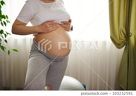 Close-up pregnant woman with naked belly, practicing yoga tree pose at home. Expectant female showing her tummy with stretch marks. Skin care. Body positivity. Pregnancy. Childbirth. Healthy lifestyle 103232591