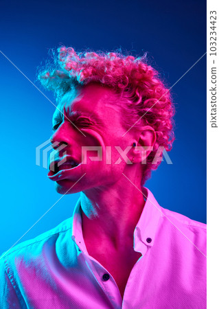 Funny image of man with curly hair posing with blowing wind on face against blue studio background in pink neon light Funny image of man with curly hair posing with blowing wind on face against blue studio background in pink neon light 103234423