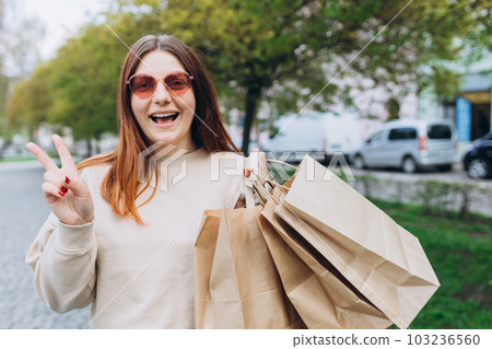 Happy young redhead woman holding shopping paper bags in hands, walking at city street, black friday. Urban lifestyle concept. Shopping banner, for mockup Happy young redhead woman holding shopping paper bags in hands, walking at city street, black friday. Urban lifestyle concept. Shopping banner, for mockup 103236560
