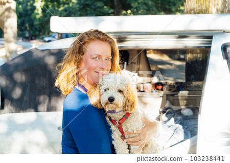 Portrait of middle-aged woman hugging her cockapoo puppy pet while standing near van camper car vehicle during road trip. Best friend. Enjoying free lifestyle, vacation, freedom. Travel with pets. 103238441