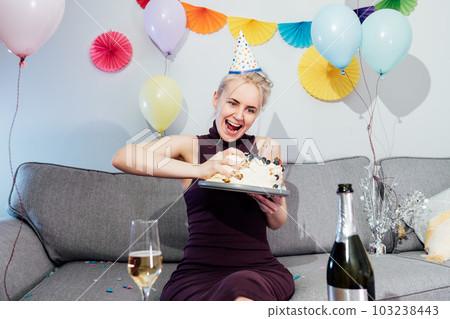 Woman in festive dress and party cap eating birthday cake with her hands, her face stained cream. A woman stuffing her face with cake. Crazy celebrating birthday party at home. Selective focus. Woman in festive dress and party cap eating birthday cake with her hands, her face stained cream. A woman stuffing her face with cake. Crazy celebrating birthday party at home. Selective focus. 103238443