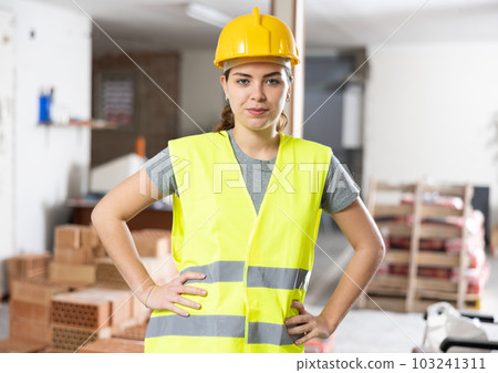 Confident female builder in yellow vest and hard hat at construction site 103241311