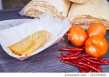 Pork tamale jalapeno and Roma tomatoes filling in corn husk on white sheet with nearby ingredients Pork tamale jalapeno and Roma tomatoes filling in corn husk on white sheet with nearby ingredients 103244895