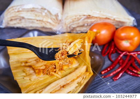 Open tamale and fork full of pork filling background red peppers, tomatoes and corn husks background Open tamale and fork full of pork filling background red peppers, tomatoes and corn husks background 103244977
