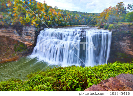 Stunning waterfall pouring over cliffs with drops splashing on lens and fall forest around 103245416