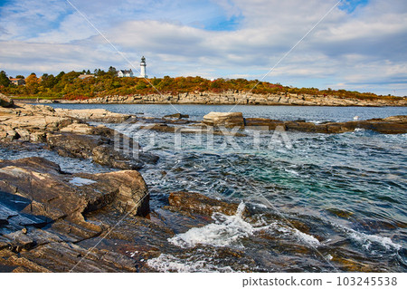 Maine coastline with waves crashing over shallow rocks and lighthouse in background Maine coastline with waves crashing over shallow rocks and lighthouse in background 103245538