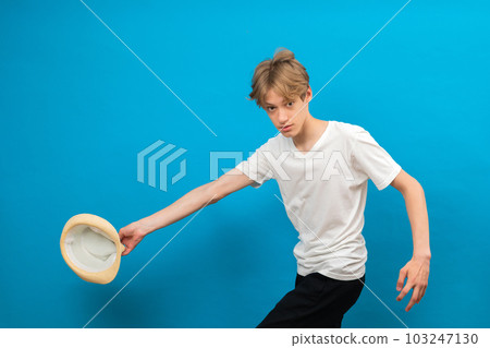 Cheerful energetic teenager boy running and holding white hat in hand in studio on blue background Cheerful energetic teenager boy running and holding white hat in hand in studio on blue background 103247130