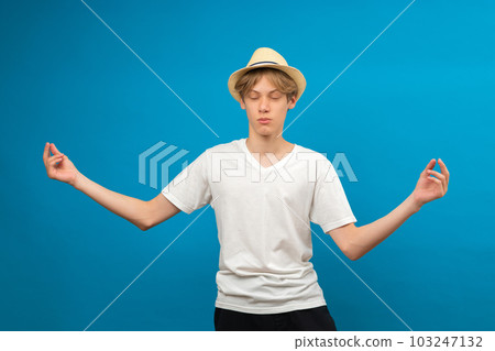 Zen. Relaxed caucasian teen boy meditating with closed eyes over blue background, calm caucasian male teen practicing yoga, keeping hands in open gesture while posing in studio, copy space 103247132