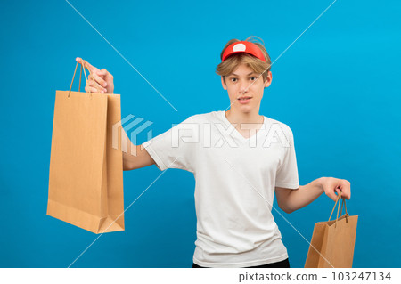 Young seller in casual white t-shirt with paper bag isolated on blue background studio portrait. People lifestyle concept. Mock up copy space. Teen holding package bag with purchases Young seller in casual white t-shirt with paper bag isolated on blue background studio portrait. People lifestyle concept. Mock up copy space. Teen holding package bag with purchases 103247134