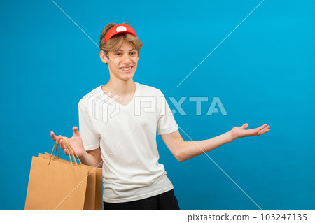 Young seller in casual white t-shirt with paper bag isolated on blue background studio portrait. People lifestyle concept. Mock up copy space. Teen holding package bag with purchases Young seller in casual white t-shirt with paper bag isolated on blue background studio portrait. People lifestyle concept. Mock up copy space. Teen holding package bag with purchases 103247135