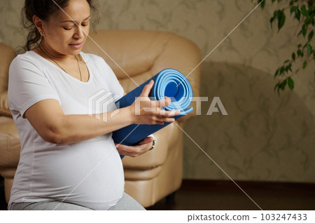Beautiful multi-ethnic pregnant expectant woman in white t-shirt, holding blue exercise mat, ready for fitness and pregnancy yoga practice at home. Healthy active lifestyle. Maternity. Expecting baby 103247433