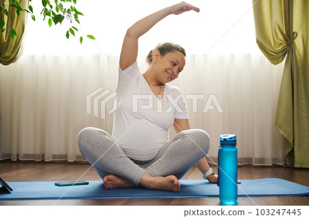 Smiling positive pregnant woman practicing pregnancy yoga, repeating after coach online, outstretching right hand, sitting in lotus pose on a blue exercise mat with digital gadget. Prenatal stretching Smiling positive pregnant woman practicing pregnancy yoga, repeating after coach online, outstretching right hand, sitting in lotus pose on a blue exercise mat with digital gadget. Prenatal stretching 103247445