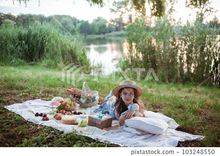 A girl in straw hat is on a picnic in a park on the banks of a river A girl in straw hat is on a picnic in a park on the banks of a river 103248550