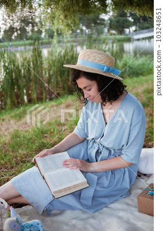 A woman in straw hat sits on a picnic in a park on the banks of a river 103248651