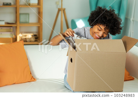African girl unpacking delivery looking in box. Happy woman opening carton box. Female getting parcel looking at delivered goods items. Satisfied client positive feedback quick express deliver 103248983