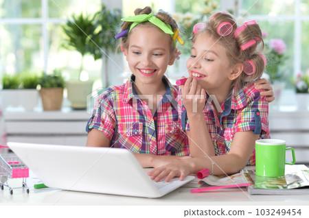 Cute twin girls in hair curlers with laptop at home appying lipstick Cute twin girls in hair curlers with laptop at home appying lipstick 103249454