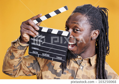 Smiling man holding cinematography clapperboard, posing confident in front of camera in studio over yellow background. African american person working in film production industry Smiling man holding cinematography clapperboard, posing confident in front of camera in studio over yellow background. African american person working in film production industry 103250068