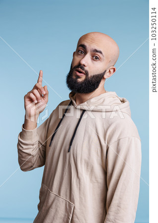 Inspired arab man having idea and raising index finger up while looking at camera. Young bald bearded person with excited facial expression pointing upwards studio portrait 103250114