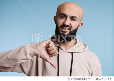 Arab man using thumb down gesture to show disapprovement while looking at camera with negative emotion grimace. Young adult person expressing disagreement studio portrait 103250233