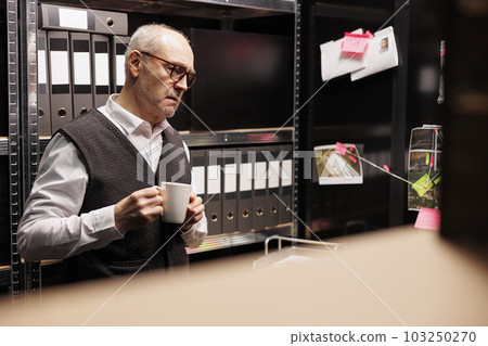 Old police officer reading legal documents in arhive room, working overhours at criminal case. Elderly private investigator analyzing crime scene evident, trying to plan startegy to catch suspect Old police officer reading legal documents in arhive room, working overhours at criminal case. Elderly private investigator analyzing crime scene evident, trying to plan startegy to catch suspect 103250270