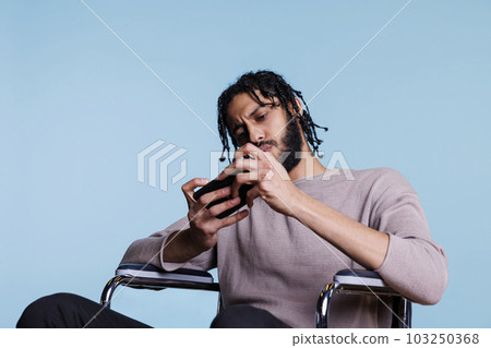 Focused arab man with disability playing video game on smartphone while sitting in wheelchair. Concentrated person with physical impairment using mobile phone application Focused arab man with disability playing video game on smartphone while sitting in wheelchair. Concentrated person with physical impairment using mobile phone application 103250368