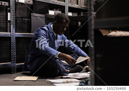 Diverse team sitting on floor in arhive room, working overtime at criminal investigation. Private detectives analyzing crime scene evidence, trying to catch mysterious suspect. Law enforcement concept Diverse team sitting on floor in arhive room, working overtime at criminal investigation. Private detectives analyzing crime scene evidence, trying to catch mysterious suspect. Law enforcement concept 103250400