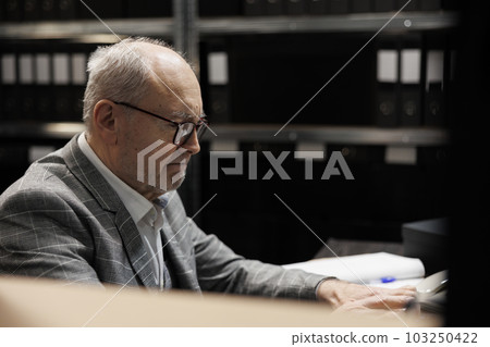 Elderly bookkeeping businessman at file cabinet office desk checking accounting analytical data paperwork. Administration senior executive in business company repository filled with document folders Elderly bookkeeping businessman at file cabinet office desk checking accounting analytical data paperwork. Administration senior executive in business company repository filled with document folders 103250422