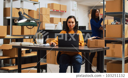 Asian owner doing supplies logistics with laptop, writing inventory information on files for startup business development. Entrepreneur planning products shipment in storage room. 103251132
