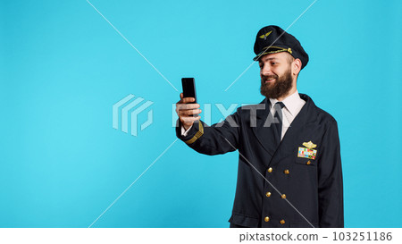 Professional airliner answering video call meeting on mobile phone, attending online remote teleconference with webcam in studio. Young airplane captain talking on videoconference chat. 103251186