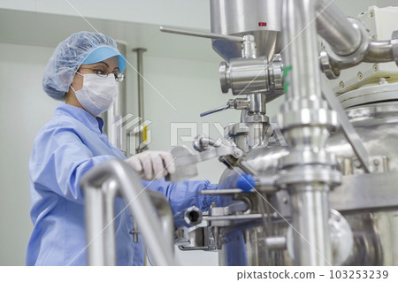 Pharmaceutical Female Worker Wearing Protective Clothing in Sterile Environment and Standing Next to Manufacturing Machine at Pharmaceutical Plant 103253239