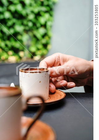 A woman takes a cup of coffee, Turkish black frothy coffee on a black stone table with branches of wild ivy in the background. Vertical frame, selective focus A woman takes a cup of coffee, Turkish black frothy coffee on a black stone table with branches of wild ivy in the background. Vertical frame, selective focus 103253805