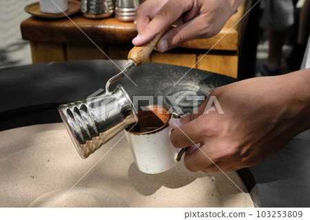 Barista pours freshly made Turkish coffee on the sand in a cezve into a cup, close-up. A professional barista prepares coffee by hand in the traditional way. Selective focus, space for text 103253809