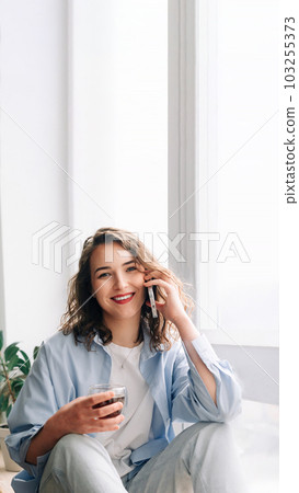 Happy woman chatting on phone with sister or friend over tea on kitchen windowsill. Pleasant 103255373
