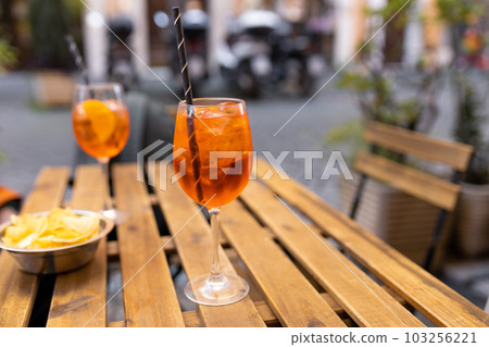 Aperol Spritz Cocktail. Alcoholic beverage based on table with ice cubes and oranges outdoors. Served cocktail with orange slice and straw placed on wooden table of sidewalk cafe in Italy 103256221