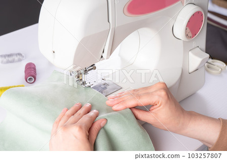 Female hands stitching white fabric on modern sewing machine at workplace in atelier Female hands stitching white fabric on modern sewing machine at workplace in atelier 103257807