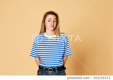Portrait of young girl in striped shirt standing with doubtful face against yellow studio background. Misunderstanding Portrait of young girl in striped shirt standing with doubtful face against yellow studio background. Misunderstanding 103258151