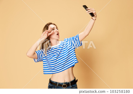Portrait of young girl in stripes shirt taking selfie with mobile phone against yellow studio background 103258180